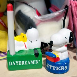 A green ceramic pen holder featuring Snoopy lying down with Woodstock and a red-and-white pen holder that reads "DAYDREAMIN'", set next to a blue paperweight featuring Snoopy at a typewriter that reads "LOVE LETTERS" on the base.