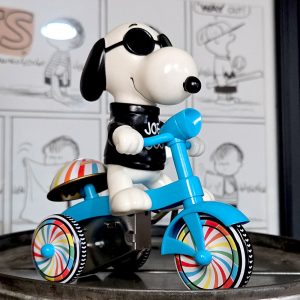 A plastic toy figure of Joe Cool Snoopy wearing sunglasses and a black "JOE COOL" sweatshirt, riding a bright blue tricycle with rainbow-swirled wheels, set against a blurred background showing Peanuts comic strip panels.