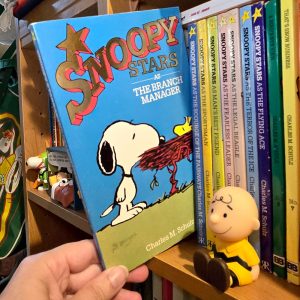 A person holding the Peanuts paperback book Snoopy Stars as the Branch Manager by Charles M. Schulz, with Snoopy and Woodstock on the cover. Next to it sits a small Charlie Brown figurine in front of a shelf filled with other Peanuts books.