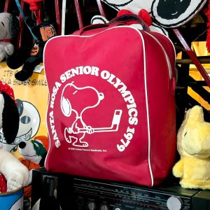 A red zippered vinyl bag with white piping and handles, printed with a white graphic of Snoopy playing ice hockey encircled by the text "Santa Rosa Senior Olympics 1979" and "©1958 United Feature Syndicate, Inc." The bag sits atop a vintage stereo receiver surrounded by various Peanuts plush toys, including a yellow Woodstock.