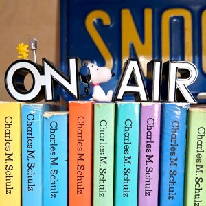 A decorative black "ON AIR" sign sitting atop a row of colorful Peanuts books by Charles M. Schulz. Small figurines of Snoopy wearing blue headphones and Woodstock sitting on a microphone are perched on top of the letters.
