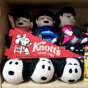 A red felt Knott's Berry Farm pennant banner featuring Snoopy in a cowboy hat leaning on a fence, placed in front of three Lucy character dolls sitting on a shelf. Below the pennant, three plush Snoopy head balls are lined up in a row.