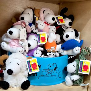 A wooden shelf filled with various Snoopy plush toys, several with Peanuts tags attached. In the center is a blue plastic container printed with a black illustration of Snoopy holding a dog dish. In the foreground, a fluffy white Snoopy plush as a sheep on the left, and a Snoopy plush wearing a green snake costume sits on the right.