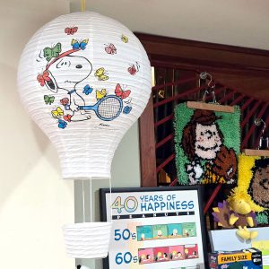 A white paper hot air balloon lantern featuring Snoopy holding a tennis racket surrounded by colorful butterflies. Below it sits a framed poster reading "40 Years of Happiness Peanuts", along with a framed Charlie Brown latch-hook craft and a plush Woodstock in the background.