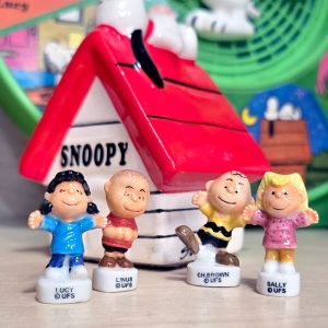Four king cake trinket Peanuts character figurines standing in a row in front of a white ceramic Snoopy doghouse bank with a red roof. From left to right: Lucy in a blue dress, Linus in a red shirt, Charlie Brown balancing on one foot, and Sally in a pink dress, each with their name printed on the white base.