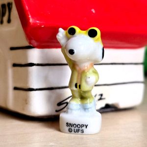 A king cake trinket of Snoopy wearing lounge suit, standing with one hand raised on a white base reading "SNOOPY © UFS". In the background, Snoopy's red-roofed doghouse is softly out of focus.