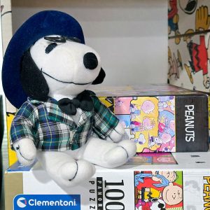 A plush Snoopy doll dressed in a scholar or professor outfit featuring a dark blue hat, black frame glasses, a black bow tie, and a green and blue plaid jacket. He sits on top of stacked Clementoni Peanuts puzzle boxes.