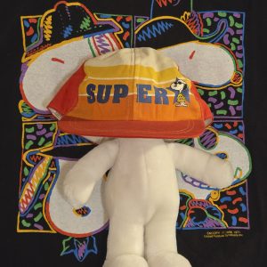 A Snoopy plush toy wearing a large, colorful neon-patterned baseball cap with the word "SUPER" across the front, displayed against a dark patterned background.