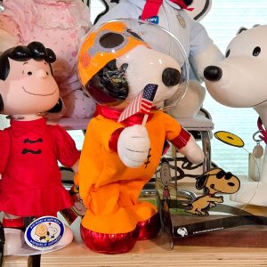 A collection of Peanuts merchandise displayed on a shelf, featuring a Lucy character doll in a red dress on the left, an Astronaut Snoopy plush in an orange spacesuit holding a small American flag in the center, and a white Snoopy plush on the right.
