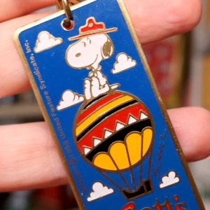 Vintage Snoopy hot air balloon key chain with a yellow and orange balloon, clouds, and text details against a hand background