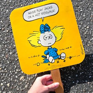 A hand holds a vintage yellow Peanuts fan against a dark asphalt background. The fan features Sally sitting on the ground wearing a blue dress and hairbow, surrounded by jacks, with a speech bubble that reads, "Never play jacks on a hot sidewalk!"