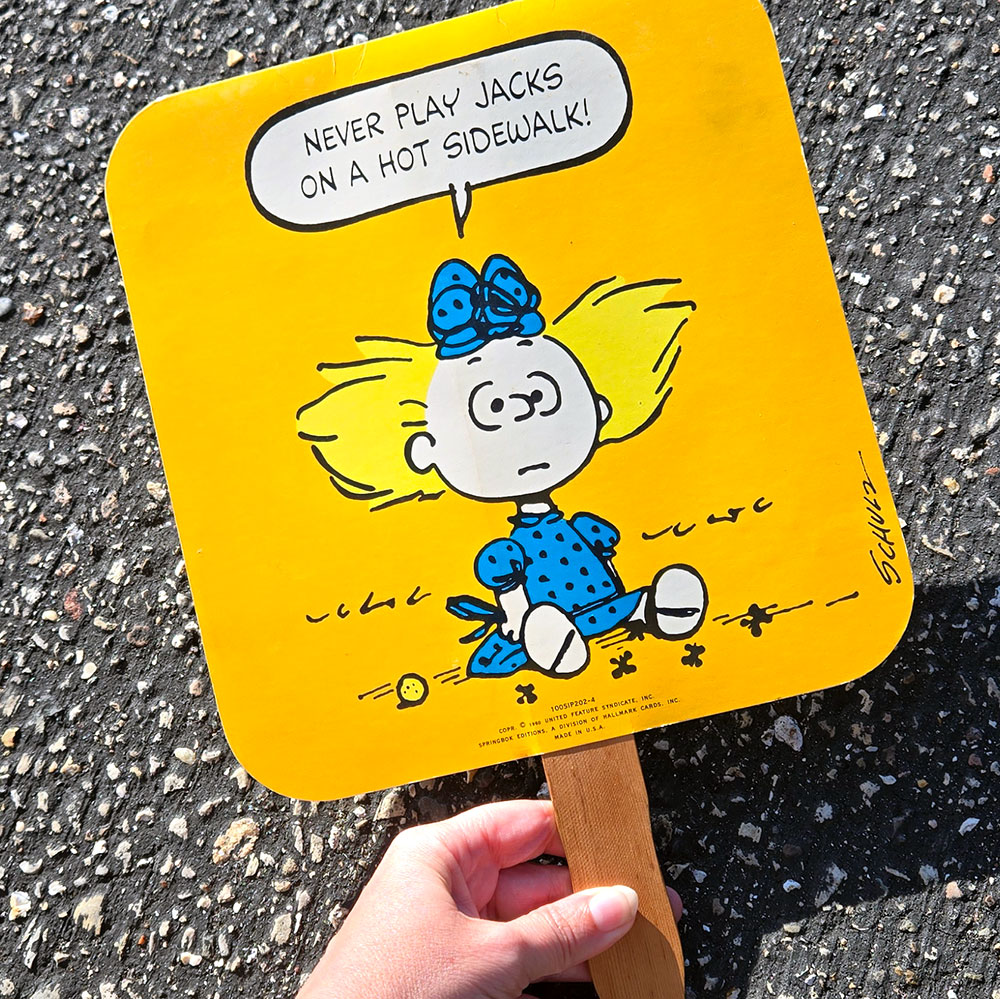 A hand holds a vintage yellow Peanuts fan against a dark asphalt background. The fan features Sally sitting on the ground wearing a blue dress and hairbow, surrounded by jacks, with a speech bubble that reads, "Never play jacks on a hot sidewalk!"