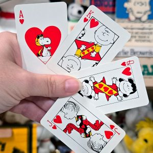 A hand holding four Peanuts playing cards featuring hearts, including an Ace with Snoopy inside the heart pip, a King with Charlie Brown, a Jack Linus, and Queen Lucy.