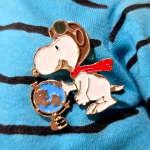 Snoopy Flying Ace enamel pin wearing his aviator helmet and a red scarf, looking down at a small blue and bronze globe resting on a stand.