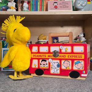 A large yellow Woodstock plush toy with spiked hair standing next to a wooden red "Peanuts Museum and Express" double-decker bus toy featuring illustrations of Charlie Brown, Snoopy, and friends.