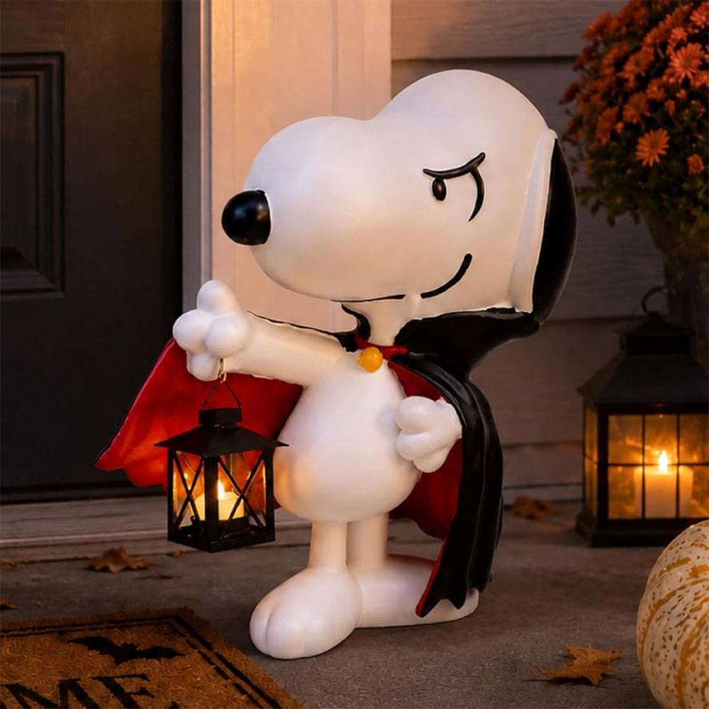 Snoopy dressed in a vampire cape holding a lit lantern on a porch with autumn decor.