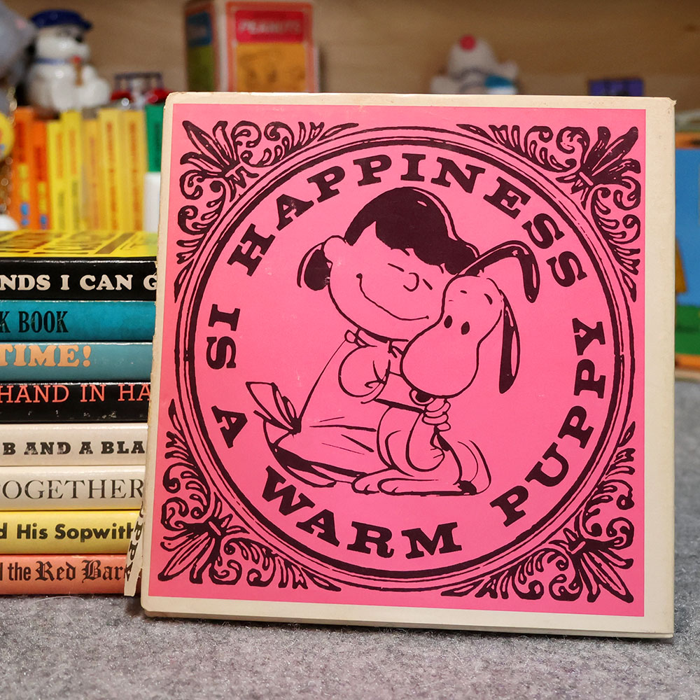 A close-up of the vintage book Happiness Is a Warm Puppy featuring a bright pink square cover with ornate black floral borders. Inside a circular frame, a smiling Lucy rests his cheek against Snoopy, surrounded by the bold text "HAPPINESS IS A WARM PUPPY." Other stacked books are visible in the blurred background.