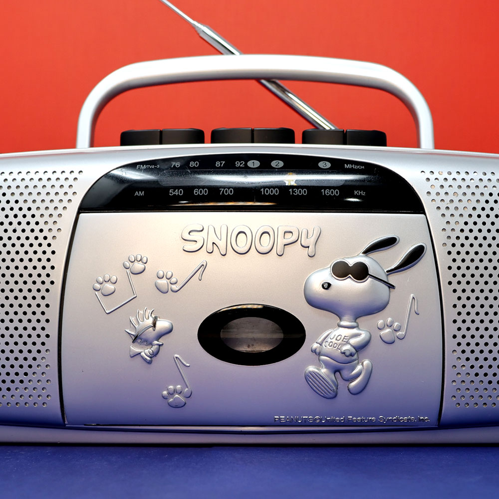 Vintage silver Snoopy AM/FM radio with a handle, featuring an illustration of Joe Cool Snoopy wearing sunglasses and musical notes on the front.