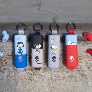 Four Skullcandy wireless earbud charging cases decorated with Peanuts graphics, lined up next to loose blue and red earbuds on a textured stone surface.