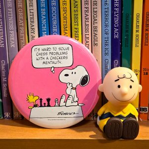 A pink round Peanuts collectible plaque featuring Snoopy and Woodstock playing a game of chess, accompanied by a Charlie Brown plush doll sitting on a wooden surface, with a row of colorful book spines displayed in the blurred background.