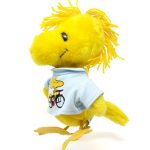 Yellow plush Woodstock stuffed bird standing on a white background, wearing a light blue doll t-shirt featured in the Snoopy's Wardrobe collection, printed with an illustration of Woodstock riding a red bicycle.