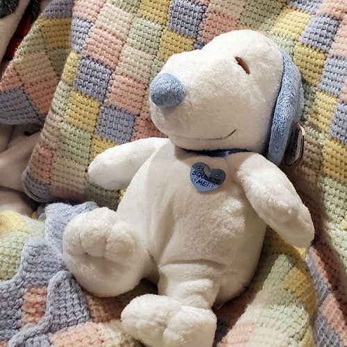 A Ty Snoopy plush toy sitting on a pastel patterned blanket, wearing a blue collar with a heart-shaped "Squeeze Me!" tag, with textured blue ears and nose.
