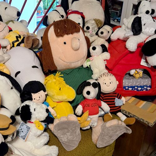 Collection of various Peanuts plush toys, including multiple Snoopy, Peppermint Patty, and Woodstock stuffed animals piled together on a couch.