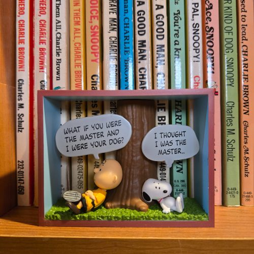 A miniature Peanuts diorama displayed in front of a row of Charles M. Schulz books, featuring Charlie Brown resting against a tree asking "WHAT IF YOU WERE THE MASTER AND I WERE YOUR DOG?" while Snoopy sits beside him thinking "I THOUGHT I WAS THE MASTER..".