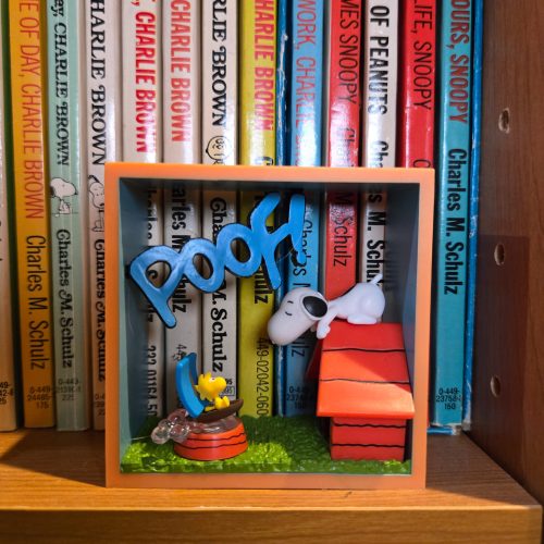 A miniature Peanuts display scene set in front of a row of Charles M. Schulz books, featuring Woodstock in a small boat on the left, Snoopy lying atop his red doghouse on the right, and a blue 3D word cutout reading "POOF!" mounted across the front.