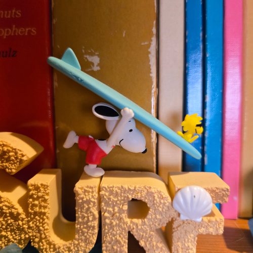 A desk display figurine featuring the word "SURF" in yellow sand-textured 3D block letters with a seashell accent on the letter "F," while Snoopy lies on a blue surfboard attached to the top alongside Woodstock.