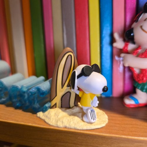 A miniature figurine of Snoopy dressed as Joe Cool, wearing a yellow shirt and black sunglasses while standing on a sandy base with his wooden surfboard, set against a bookshelf background.