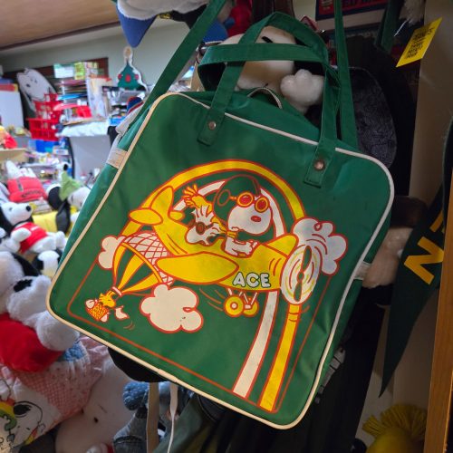 A green vintage Snoopy vinyl flight bag featuring an illustration of Snoopy as the Flying Ace wearing goggles and a scarf in an airplane marked "ACE", accompanied by Woodstock in a scarf, set against a room filled with Peanuts collectibles.