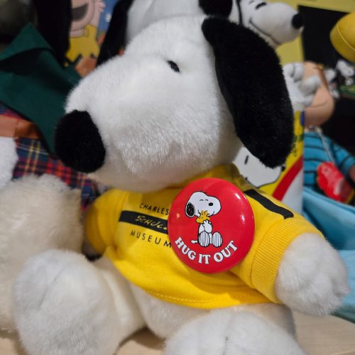 A Snoopy plush toy wearing a yellow t-shirt that reads "CHARLES M. SCHULZ MUSEUM", featuring a red pinback button pinned to its chest with an illustration of Snoopy and the text "HUG IT OUT".