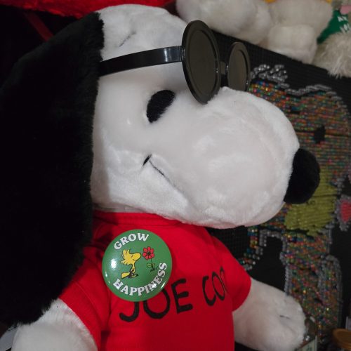 A Snoopy plush toy wearing black sunglasses and a red shirt that reads "JOE COOL", with a green pinback button pinned to its chest featuring Woodstock and the words "GROW HAPPINESS".