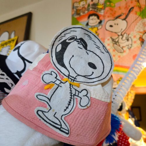A close-up view of a pink and white fabric hat featuring an embroidered patch of Astronaut Snoopy wearing a bubble space helmet and suit, set against a background filled with Peanuts collectibles.