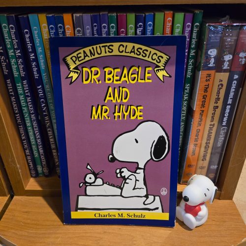 A paperback book titled "Peanuts Classics: Dr. Beagle and Mr. Hyde" by Charles M. Schulz, displaying an illustration of Snoopy typing at his typewriter while perched on top of a giant Snoopy head, set against a bookshelf filled with Peanuts books.