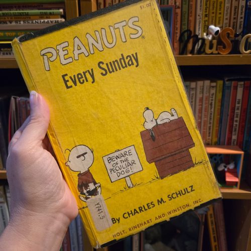A hand holding a vintage yellow library-bound edition of the Peanuts book "Peanuts Every Sunday" by Charles M. Schulz, published by Holt, Rinehart and Winston, set against a background of bookshelves filled with Peanuts collectibles.