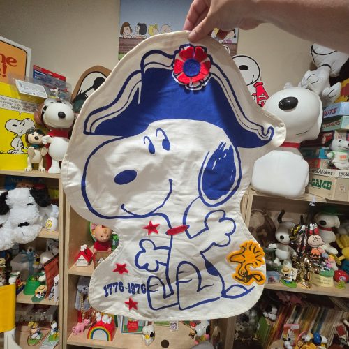 A hand holding up a vintage fabric Snoopy Bicentennial pajama bag featuring Snoopy dressed in Revolutionary War attire with a blue hat and red bow tie, displaying the dates "1776-1976" and a felt Woodstock attached near his feet, set against shelves filled with Peanuts collectibles.