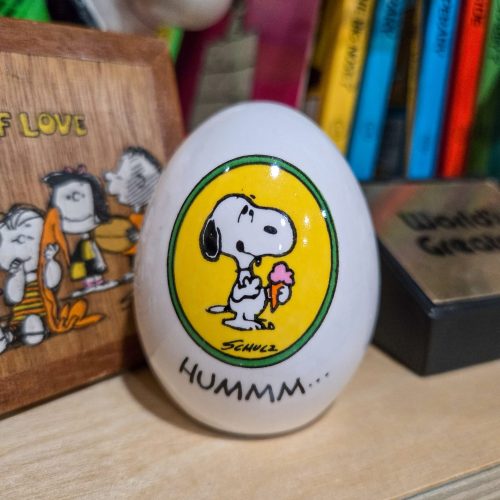 A decorative white ceramic egg featuring an illustration of Snoopy holding a pink ice cream cone against a yellow circular background bordered in green, with Schulz's signature below and the text "HUMMM..." printed near the bottom.