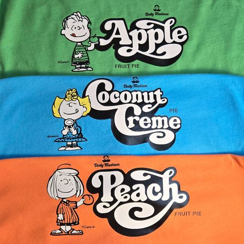 Three vintage Dolly Madison Fruit Pie T-shirts stacked on top of each other in green, blue, and orange, featuring Linus next to "Apple," Sally next to "Coconut Creme," and Peppermint Patty next to "Peach."