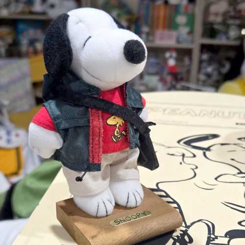 Plush Snoopy doll standing on a wooden base labeled "SNOOPY," dressed in a red sweatshirt featuring a Woodstock graphic, a dark denim jacket with white lining, and a black cross-body strap.