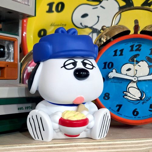 A plastic figurine of Olaf wearing a blue cap, sitting down and holding a red bowl of yellow food with a bone resting on top. In the background are a blue and a yellow vintage Peanuts alarm clock featuring Snoopy.