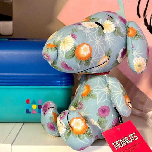 A plush Snoopy stuffed animal crafted from light blue fabric decorated with colorful orange, pink, and white floral and geometric patterns, featuring a red neck ribbon and a red Peanuts tag.