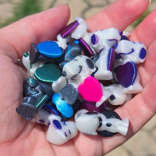 A person's open hand holding a pile of small 3D-printed plastic Snoopy figures in various colors, including white, metallic blue, green, black, purple, and a bright pink heart.