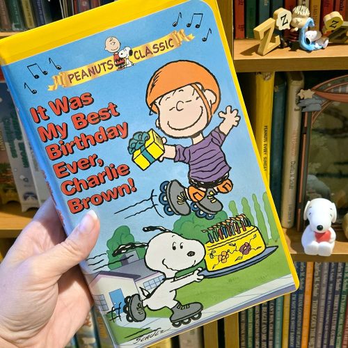 Cover of It Was My Best Birthday Ever, Charlie Brown! VHS Tape against a bright blue background, featuring Linus wearing an orange hat and purple striped shirt while wearing roller skates and holding a yellow gift box, with Snoopy on roller skates and a yellow cake with lit candles at the bottom, alongside a hand holding the book in front of a bookshelf filled with books.