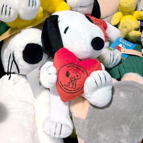 Snoopy plush toy resting among a pile of other plush items and toys, featuring Snoopy's classic black and white design with floppy black ears, embroidered facial features, whiskers, and holding a small red heart.