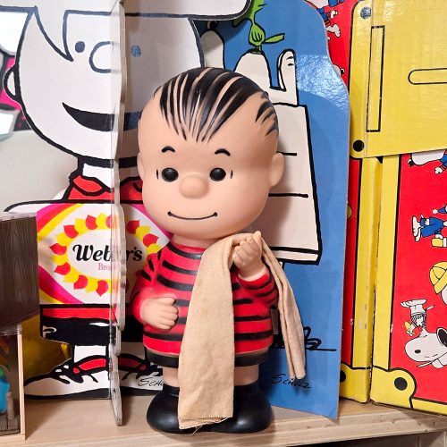 Linus van Pelt plastic Hungerford doll holding his signature security blanket, positioned in front of a Charlie Brown display and a colorful blue-and-yellow background.