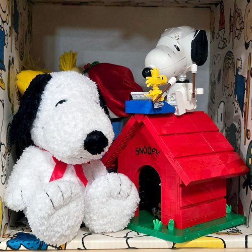 A fluffy white Snoopy plush toy sitting next to a red LEGO doghouse, on top of which a LEGO Snoopy figure and a yellow Woodstock figure are seated, all positioned inside a display shelf lined with Peanuts comic strip wallpaper.