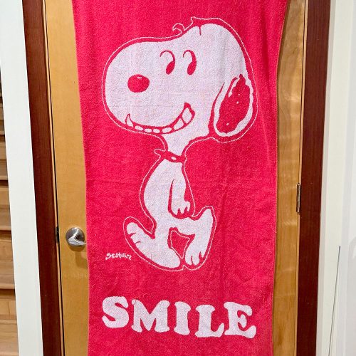 A bright red Peanuts beach towel hanging over a wooden door, featuring a large white outline illustration of Snoopy smiling with the word "SMILE" printed in bold white letters across the bottom.