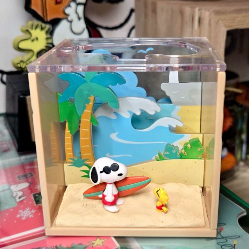 A small clear acrylic display box featuring a tropical beach diorama with Snoopy and Woodstock. Snoopy wears sunglasses and red swim trunks while holding an orange-and-teal surfboard on the sandy beach next to Woodstock. The background features layered cutout palm trees, ocean waves, and a yellow sky.
